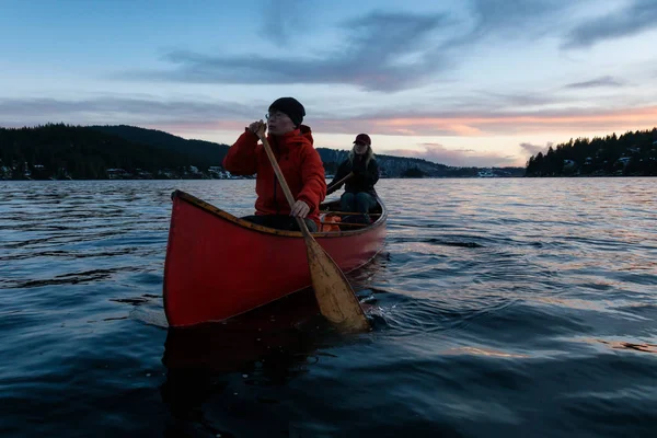 Couple friends on a wooden canoe are paddling in an inlet surrounded by Canadian mountains during a vibrant sunset. Taken in Indian Arm, near Deep Cove, North Vancouver, British Columbia, Canada.