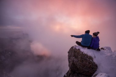 Sırasında canlı bir gün batımı manzarasına bakan sarp bir uçurum kenarında oturan genç bir çift. Üstünde tepe-in St Mark's Peak, Kuzey Vancouver, Bc, Kanada alınan.