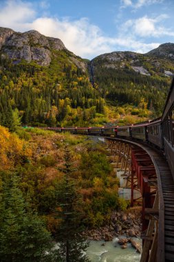 Skagway, Alaska, ABD - 24 Eylül 2019: Eski Tarihi Demiryolu Treni bulutlu bir yaz sabahı turistlerle birlikte Beyaz Geçit 'teki ahşap bir köprüden geçiyor.