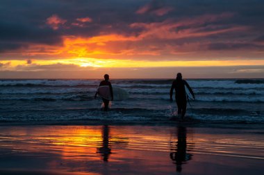Dramatik bir yaz günbatımında kumlu sahilde okyanusa doğru yürüyen sörfçüler. Seaside, Oregon, Amerika Birleşik Devletleri 'nde çekildi..