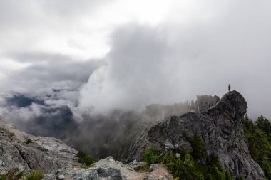 Bulutlu bir yaz sabahı engebeli bir kayalık dağın tepesinde duran maceraperest adam. Crown Dağı, Kuzey Vancouver, Bc, Kanada.