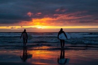 Dramatik bir yaz günbatımında kumlu sahilde okyanusa doğru yürüyen sörfçüler. Seaside, Oregon, Amerika Birleşik Devletleri 'nde çekildi..