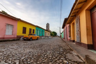 Güneşli bir gündoğumu sırasında arka planda kilisesi olan küçük bir Küba kasabasının sokaklarında eski klasik bir taksi manzarası. Trinidad, Küba.