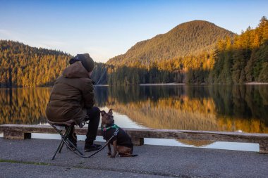 Port Moody, Vancouver, British Columbia, Canada - Feb 20, 2020: Man with a dog is fishing in the lake at White Pine Beach during a vibrant winter sunset.