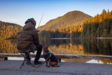 Port Moody, Vancouver, British Columbia, Canada - Feb 20, 2020: Man with a dog is fishing in the lake at White Pine Beach during a vibrant winter sunset.
