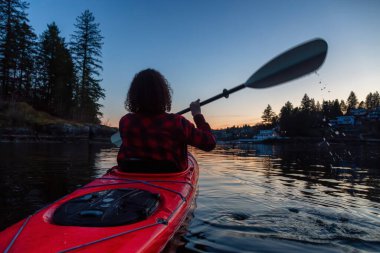 Canlı ve renkli bir günbatımında sakin okyanus suyunda Parlak Kırmızı bir kanoyla yüzen maceraperest kız. Indian Arm, Deep Cove, North Vancouver, British Columbia, Kanada 'da çekildi..