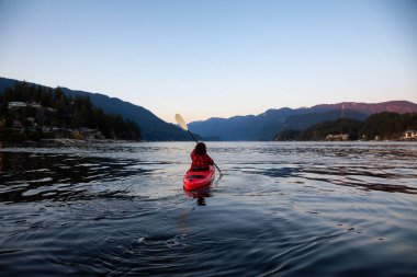 Canlı ve renkli bir günbatımında sakin okyanus suyunda Parlak Kırmızı bir kanoyla yüzen maceraperest kız. Indian Arm, Deep Cove, North Vancouver, British Columbia, Kanada 'da çekildi..