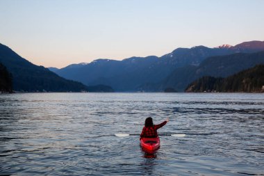 Canlı ve renkli bir günbatımında sakin okyanus suyunda Parlak Kırmızı bir kanoyla yüzen maceraperest kız. Indian Arm, Deep Cove, North Vancouver, British Columbia, Kanada 'da çekildi..