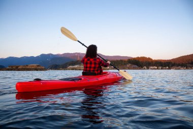 Canlı ve renkli bir günbatımında sakin okyanus suyunda Parlak Kırmızı bir kanoyla yüzen maceraperest kız. Indian Arm, Deep Cove, North Vancouver, British Columbia, Kanada 'da çekildi..