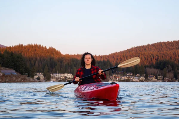 Adventurous Girl Paddling on a Bright Red Kayak in calm ocean water during a vibrant and colorful sunset. Taken in Indian Arm, Deep Cove, North Vancouver, British Columbia, Canada.