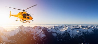 Güneşli bir gündoğumunda Rocky Dağları üzerinde uçan sarı helikopter.