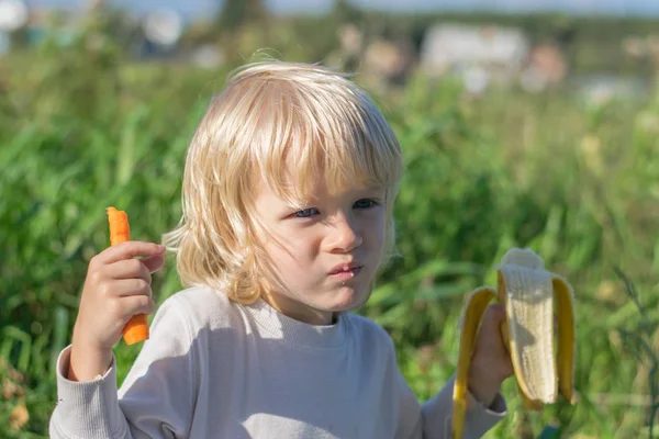 金发碧眼的小男孩吃胡萝卜和香蕉 — 图库照片