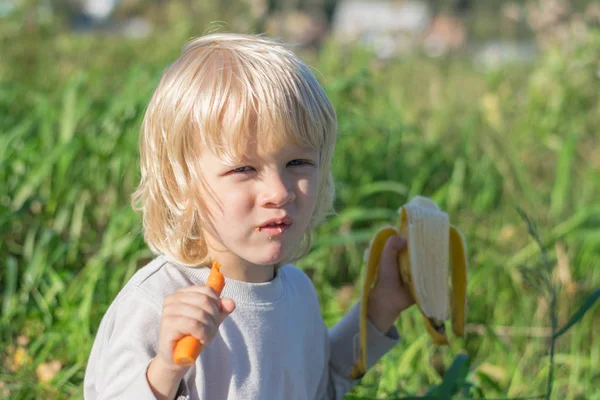 金发碧眼的小男孩吃胡萝卜和香蕉 — 图库照片