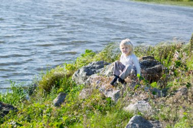 Bir kayalar nehir kıyısında oturan sarışın düşünceli çocuk