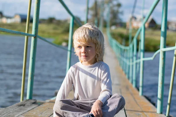 Ein glücklicher blonder Junge auf einer alten hölzernen Hängebrücke — Stockfoto