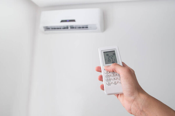 Hand holding remote control for air conditioner on white wall.