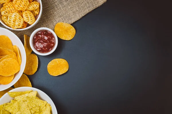 Aardappels, chips en snacks op zwarte leisteen tafel, bovenaanzicht — Stockfoto