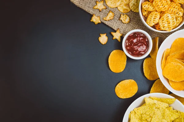 Aardappels, chips en snacks op zwarte leisteen tafel, bovenaanzicht — Stockfoto