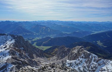 Karlı gri kayalar, aşağıdaki köy, arka planda dağlar ve bulutlu gökyüzü.