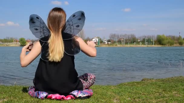Девушка снимает крылья бабочки и продолжает медитировать в позе лотоса рядом с спокойной водой — стоковое видео