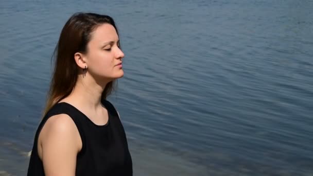 Hermosa morena con los ojos cerrados y en una camisa negra; disfrutando por la costa — Vídeos de Stock