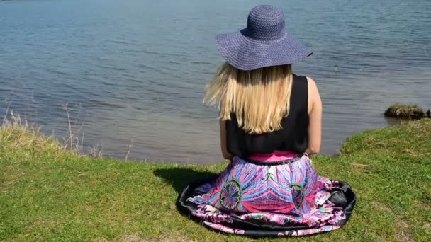 Fille avec un chapeau regarder la surface paisible de l'eau, jusqu'à ce que ses cheveux volent dans le vent — Video