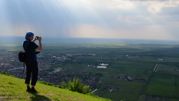 Een jonge man staande op een heuvel en fotograferen van adembenemend uitzicht op de vlakte — Stockvideo