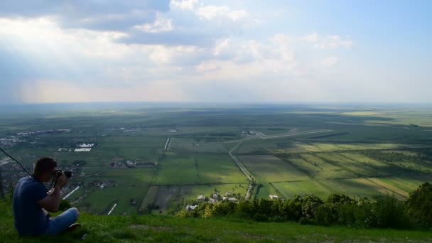 Ein junger Mann sitzt auf einem Hügel und fotografiert einen atemberaubenden Blick auf die Ebene — Stockvideo