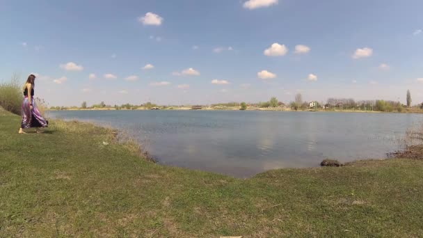 Joven mujer de pie en la orilla del río, viento soplando su pelo — Vídeo de stock