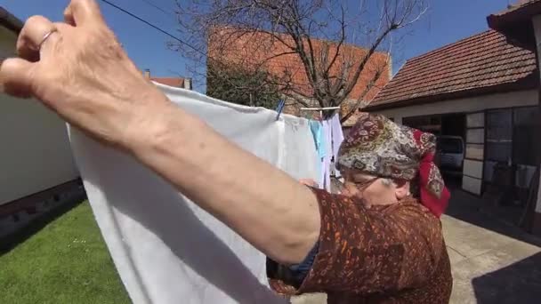 Una abuela muy vieja con una bufanda en la cabeza extendiendo la ropa a secar — Vídeos de Stock