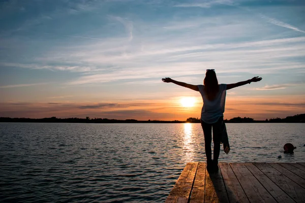 Mujer joven de pie con los brazos abiertos en el pontón de madera en el lago y viendo una colorida puesta de sol — Foto de Stock