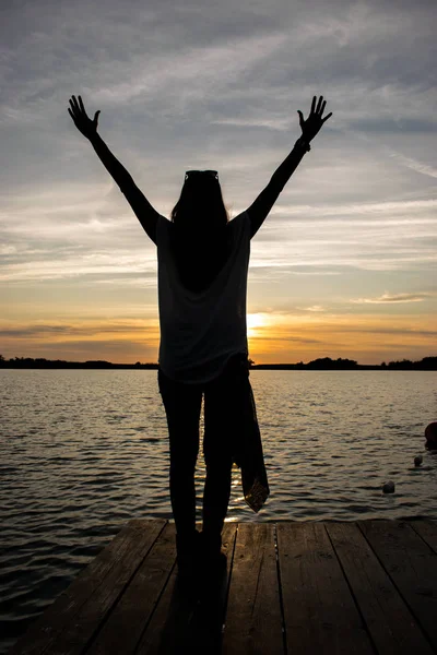 Mujer joven de pie con los brazos abiertos en el pontón de madera en el lago y viendo una colorida puesta de sol — Foto de Stock