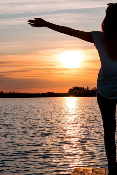 Młoda kobieta stojąc z otwartymi ramionami na ponton drewniane w jeziorze i oglądania kolorowy zachód słońca — Zdjęcie stockowe