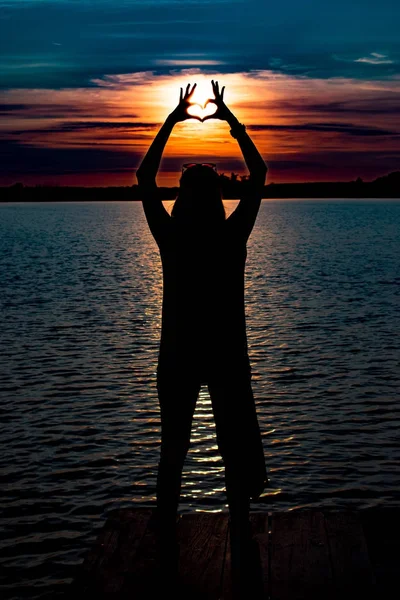 Joven mujer de pie y hace un corazón con sus manos en el pontón de madera en el lago y viendo una colorida puesta de sol — Foto de Stock