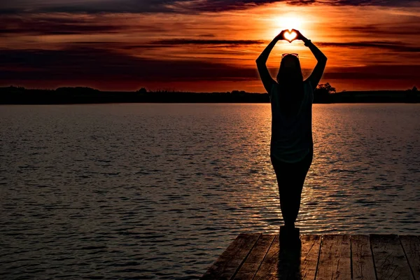 Νεαρή γυναίκα να στέκεται και να κάνει μια καρδιά με τα χέρια σε λιωμένο στο στη λίμνη και ένα πολύχρωμο ηλιοβασίλεμα — Φωτογραφία Αρχείου