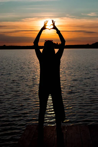 Νεαρή γυναίκα να στέκεται και να κάνει μια καρδιά με τα χέρια σε λιωμένο στο στη λίμνη και ένα πολύχρωμο ηλιοβασίλεμα — Φωτογραφία Αρχείου