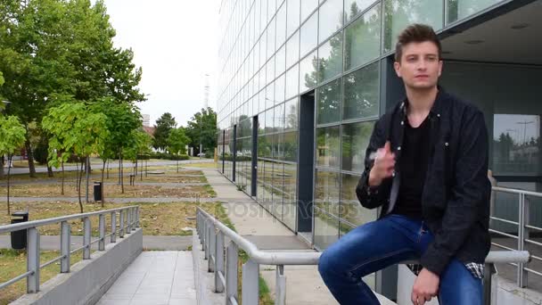 Casual genç adam bir korkuluk cam bina karşı oturup sigara içiyor — Stok video