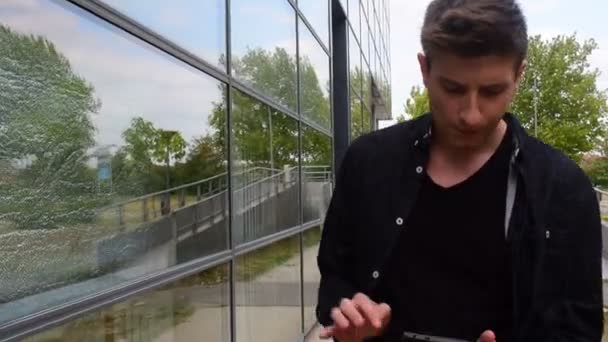 Ένας νεαρός άνδρας τον έλεγχο αλληλογραφίας, ειδήσεις και τα κοινωνικά δίκτυα στο τηλέφωνό του, περνώντας από ένα γυάλινο κτίριο — Αρχείο Βίντεο