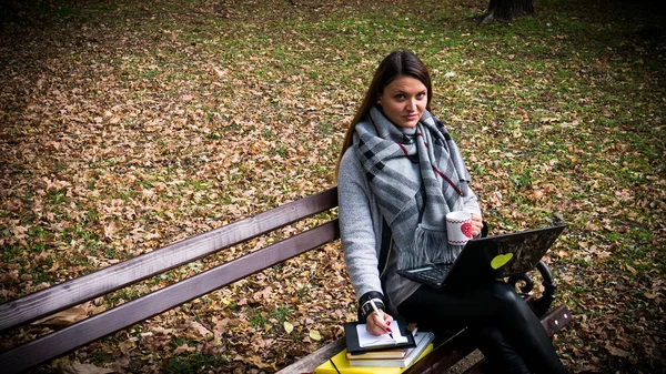 一个美丽的年轻女子坐在长凳上的公园和工作的笔记本电脑, 拿着一杯咖啡/茶, 看着相机和微笑 — 图库照片