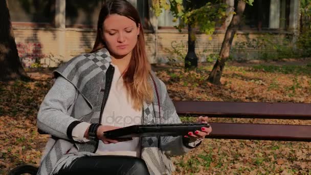 Una hermosa joven sentada en el banco en un parque y trabajando en una computadora portátil en la oficina al aire libre — Vídeo de stock