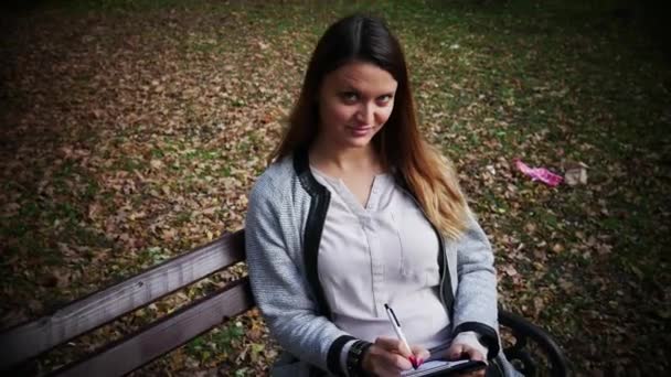 Mulher bonita está escrevendo um diário ao ar livre no parque, estudante estudando em um banco no parque, menina estudante se preparando para o exame sobre a natureza — Vídeo de Stock