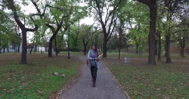 Молодая женщина в парке подходит к камере, улыбается, проходит мимо камеры и камера движется, чтобы следовать за ней — стоковое видео