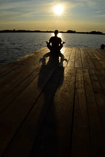Silhueta de um jovem meditando em tábuas de madeira em frente ao lago em frente ao sol — Fotografia de Stock