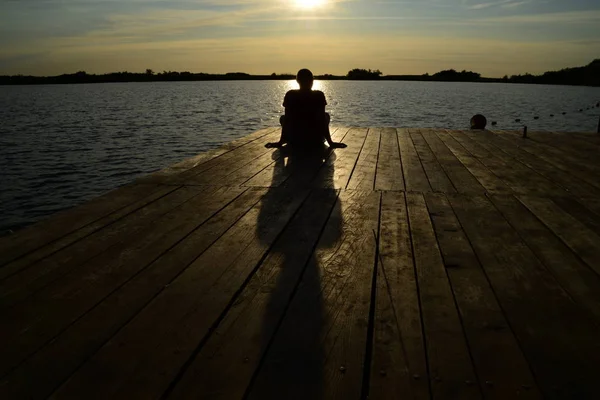 Sylwetka młodego człowieka, siedząc na jezioro o zachodzie słońca / wschód słońca — Zdjęcie stockowe