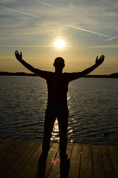 Silueta de un joven con los brazos abiertos en el aire, brazos extendidos en el aire frente al lago frente al sol — Foto de Stock