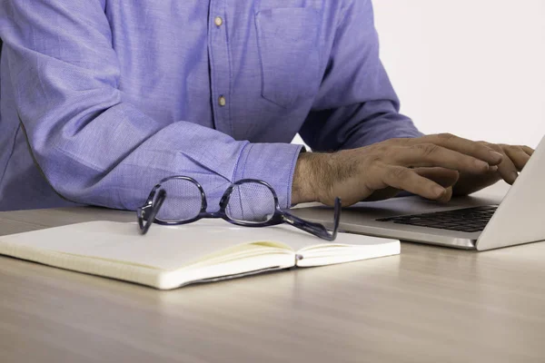 Yaşlı iş adamının elleri dizüstü bilgisayarın klavyesindeyken bilgisayarın yanındaki açık defterde gözlük takmak..