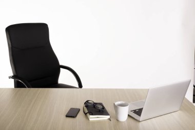 Ahşap bir masada boş siyah bir koltuk, açık bir dizüstü bilgisayar, bir fincan kahve, defter, okuma gözlüğü ve cep telefonu. Boşluk yok. Metin için alanı kopyala. 