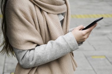 Paltolu ve fularlı bir kadın bir şehrin caddesinde elinde bir cep telefonu tutuyor. Yakın çekim..