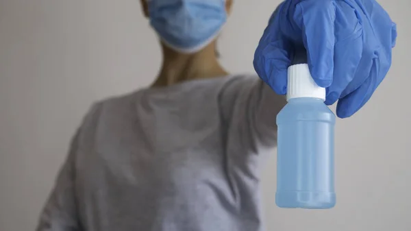 A woman with a surgical mask holds a plastic bottle of alcohol gel in her hand that is in a rubber glove