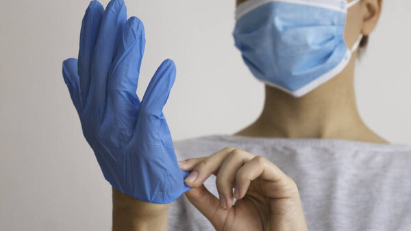 Женщина в медицинской маске надевает голубые резиновые перчатки на руки
.
